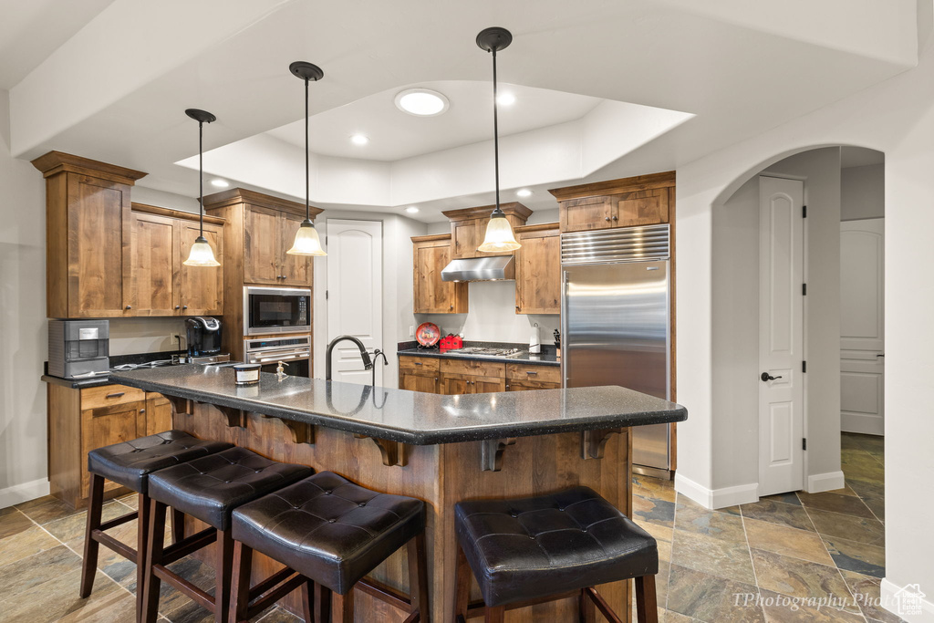 Kitchen with hanging light fixtures, a tray ceiling, extractor fan, built in appliances, and a kitchen bar