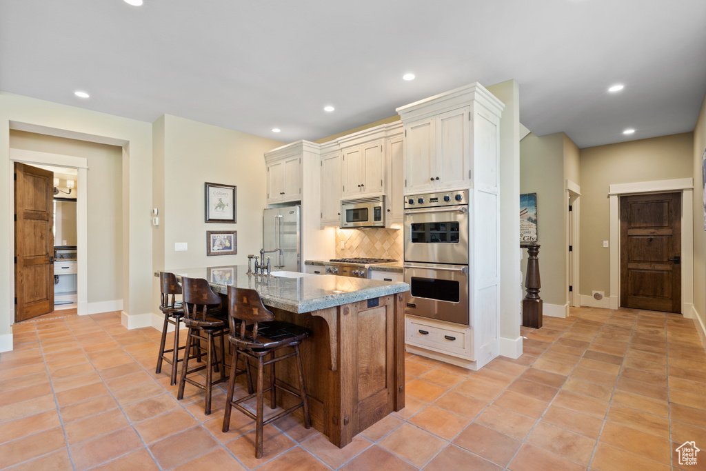 Kitchen featuring light tile patterned floors, tasteful backsplash, a kitchen bar, stainless steel appliances, and light stone counters