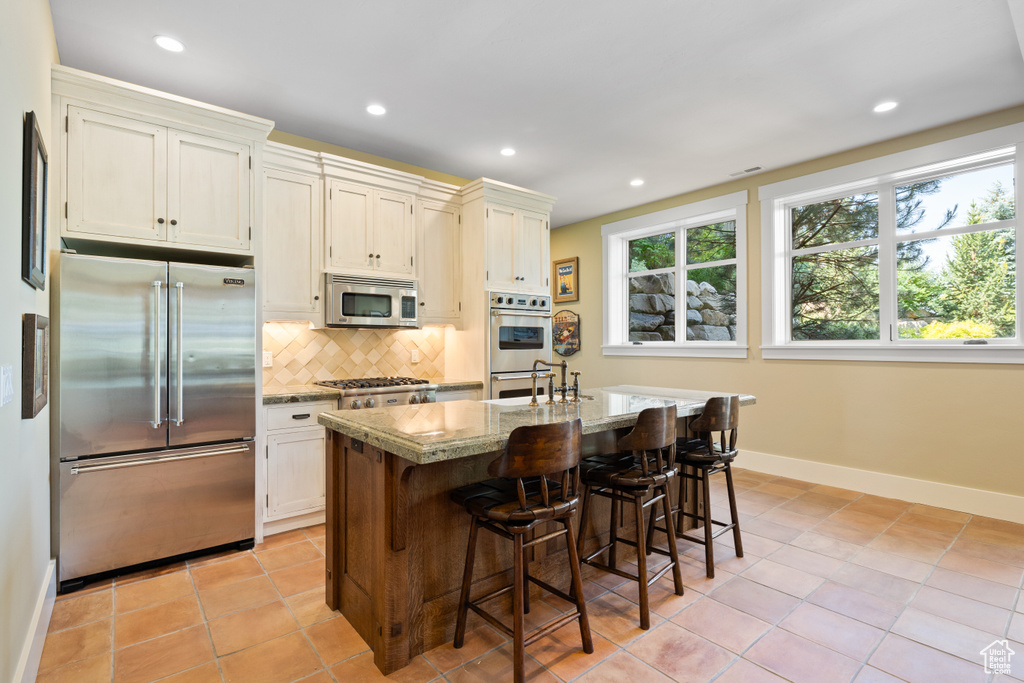 Kitchen with backsplash, an island with sink, a breakfast bar, stainless steel appliances, and light tile patterned flooring