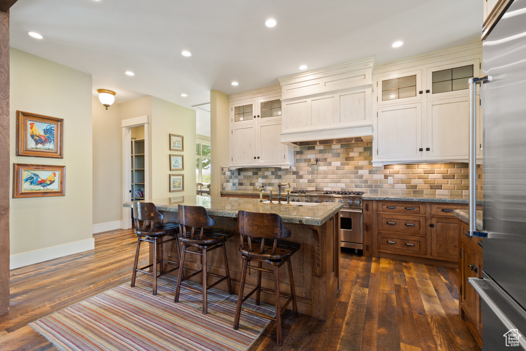 Kitchen with dark hardwood / wood-style floors, backsplash, a kitchen island with sink, light stone counters, and premium appliances