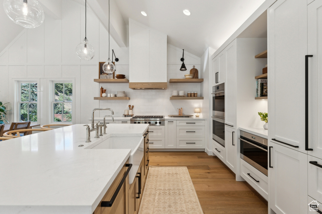 Kitchen featuring white cabinets, an island with sink, decorative light fixtures, appliances with stainless steel finishes, and light wood-type flooring