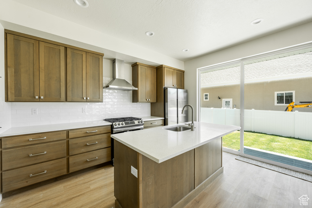 Kitchen featuring light wood-type flooring, a wealth of natural light, an island with sink, and wall chimney range hood