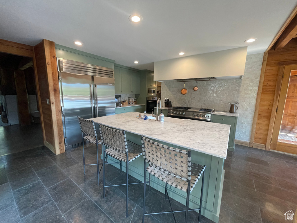 Kitchen with built in appliances, decorative backsplash, light stone counters, wall chimney range hood, and green cabinetry