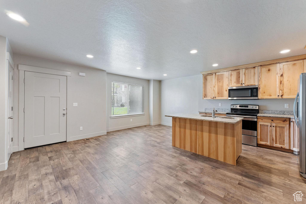 Kitchen with appliances with stainless steel finishes, light hardwood / wood-style flooring, light stone counters, and a center island with sink