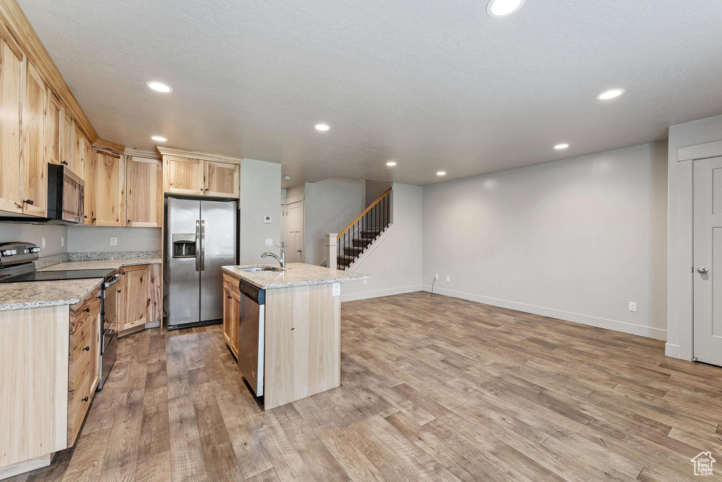 Kitchen with light stone countertops, appliances with stainless steel finishes, light hardwood / wood-style floors, an island with sink, and light brown cabinets