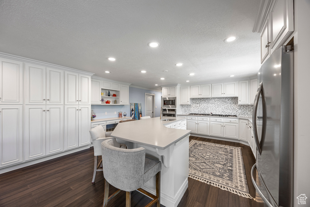 Kitchen featuring white cabinets, appliances with stainless steel finishes, dark wood-type flooring, a kitchen breakfast bar, and a kitchen island with sink