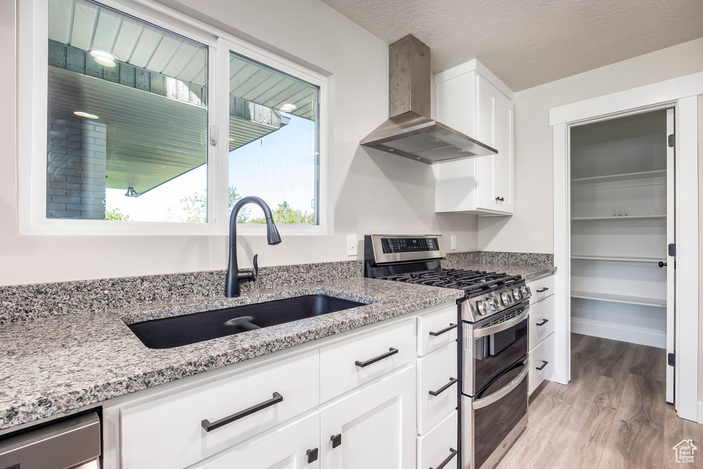 Kitchen featuring double oven range, light wood-type flooring, wall chimney range hood, sink, and white cabinets