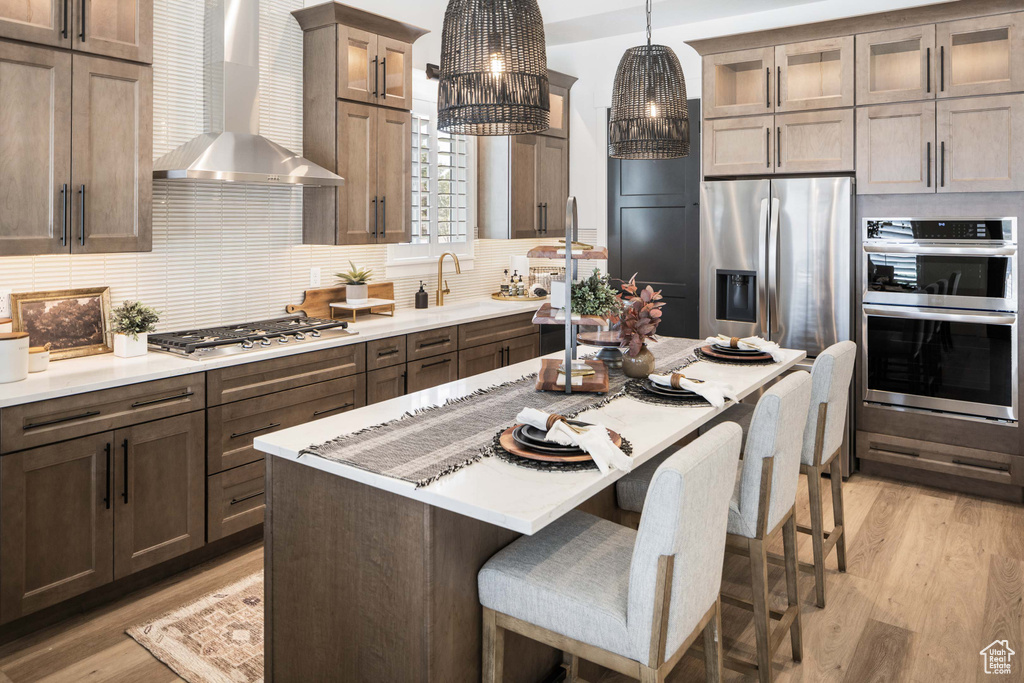 Kitchen with a kitchen island, decorative light fixtures, wall chimney exhaust hood, stainless steel appliances, and light wood-type flooring