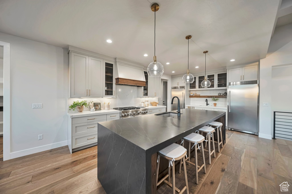 Kitchen featuring high end appliances, decorative light fixtures, a center island with sink, light hardwood / wood-style flooring, and custom range hood