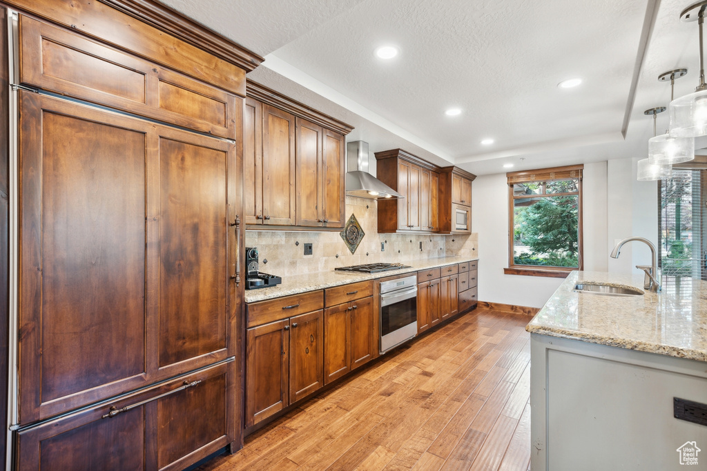 Kitchen with pendant lighting, wall chimney range hood, sink, appliances with stainless steel finishes, and light hardwood / wood-style floors