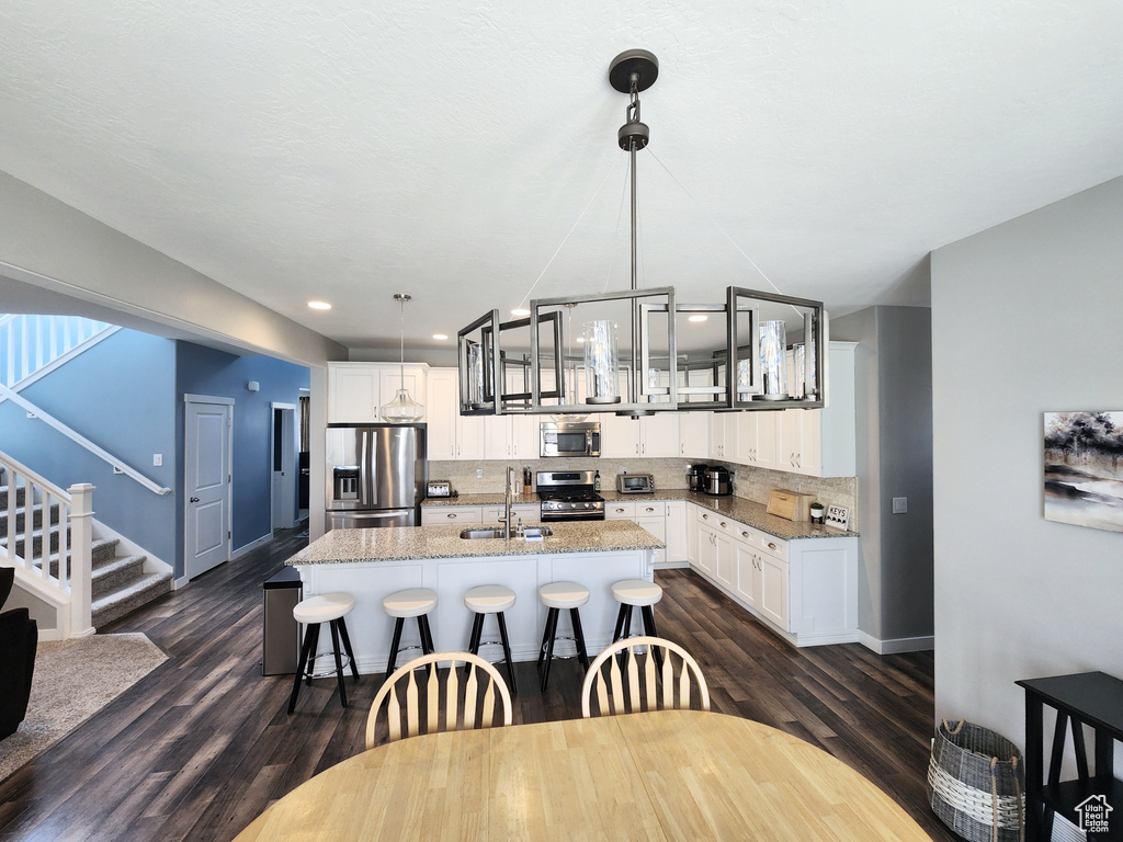 Kitchen featuring hanging light fixtures, an inviting chandelier, appliances with stainless steel finishes, dark wood-type flooring, and a kitchen island with sink