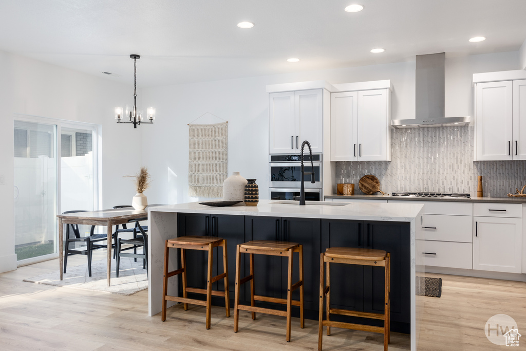 Kitchen featuring a center island with sink, wall chimney exhaust hood, decorative light fixtures, and white cabinetry