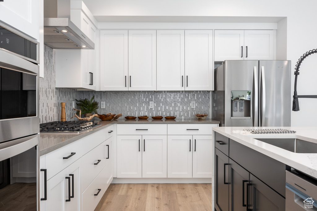 Kitchen featuring wall chimney exhaust hood, light hardwood / wood-style flooring, appliances with stainless steel finishes, white cabinetry, and light stone counters