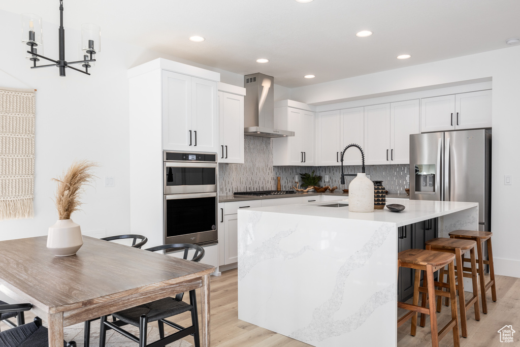 Kitchen featuring light wood-type flooring, stainless steel appliances, an island with sink, a breakfast bar, and wall chimney range hood