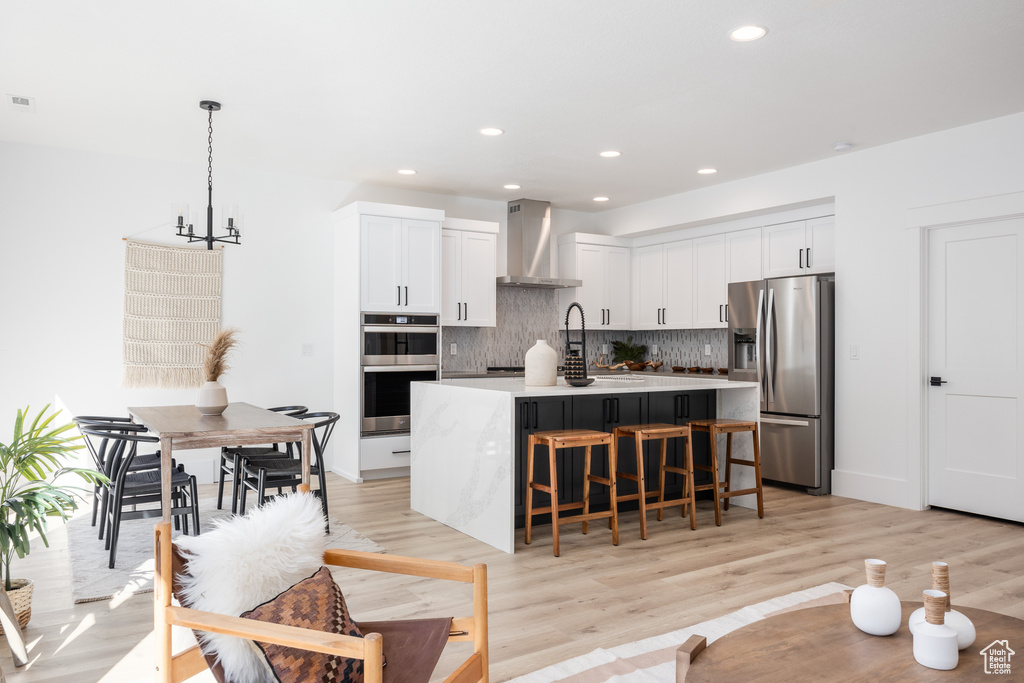 Kitchen with wall chimney range hood, a center island, appliances with stainless steel finishes, white cabinets, and light hardwood / wood-style floors