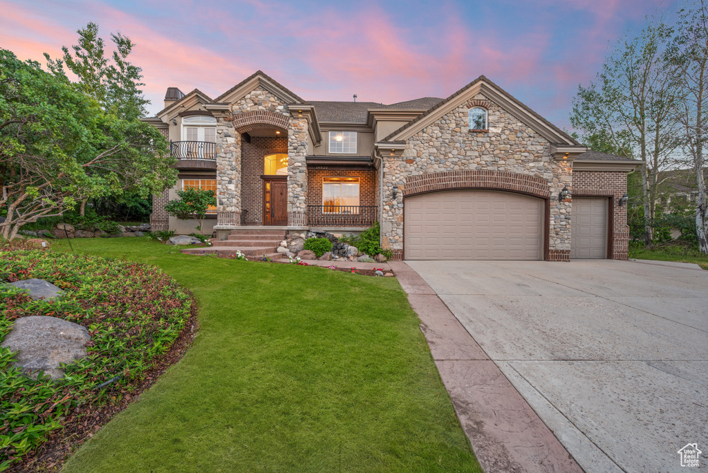 French country style house featuring a garage, driveway, a front lawn, a balcony, and stone siding