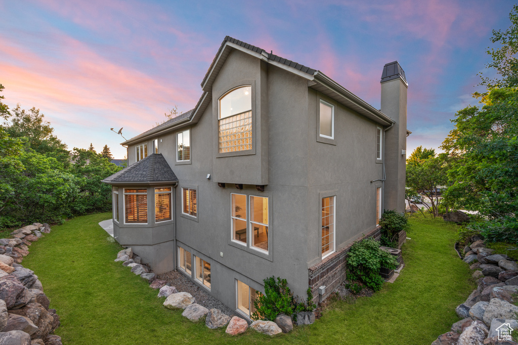 Back of house at dusk featuring stucco siding, a yard, and a chimney