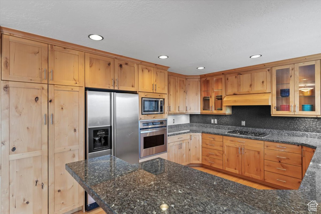 Kitchen featuring under cabinet range hood, glass insert cabinets, stainless steel appliances, and dark stone countertops