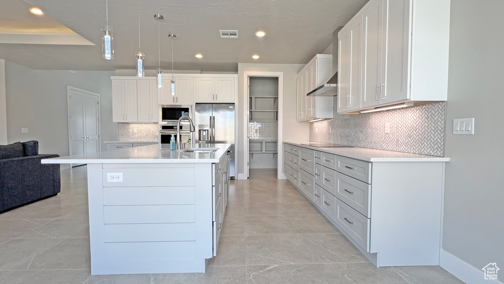 Kitchen featuring a center island with sink, appliances with stainless steel finishes, hanging light fixtures, decorative backsplash, and white cabinets