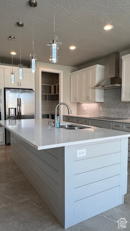 Kitchen with wall chimney range hood, decorative light fixtures, a center island with sink, and white cabinetry