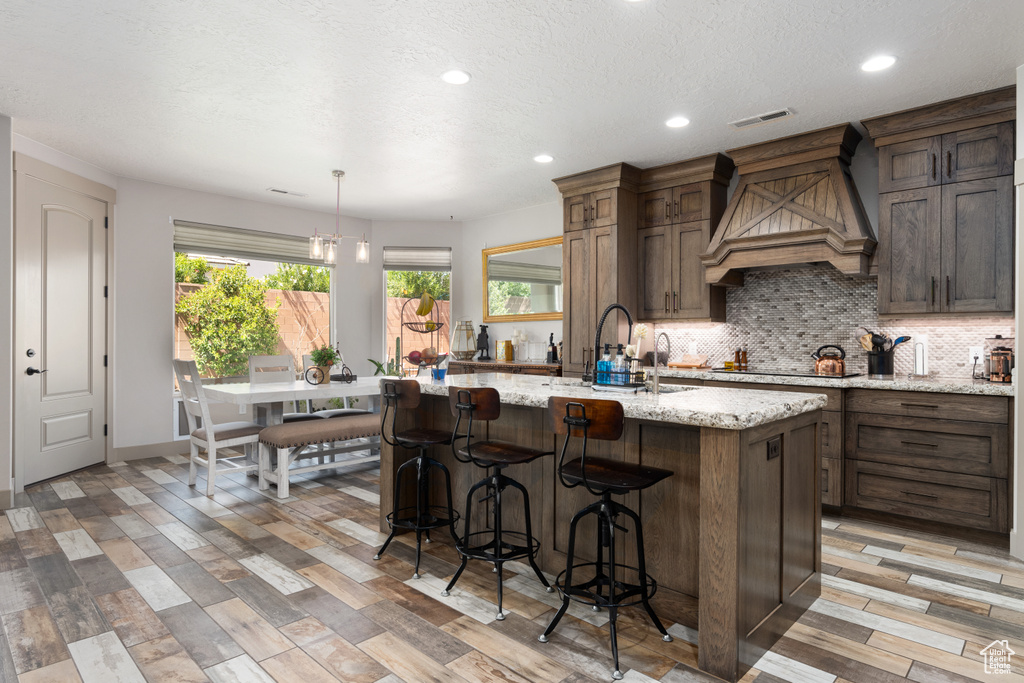 Kitchen with pendant lighting, light hardwood / wood-style flooring, premium range hood, an island with sink, and light stone countertops