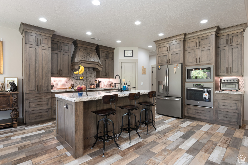 Kitchen featuring light hardwood / wood-style floors, an island with sink, a breakfast bar, stainless steel appliances, and custom exhaust hood