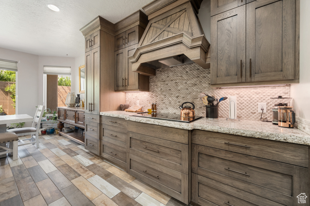 Kitchen with black electric cooktop, custom range hood, light stone counters, and backsplash