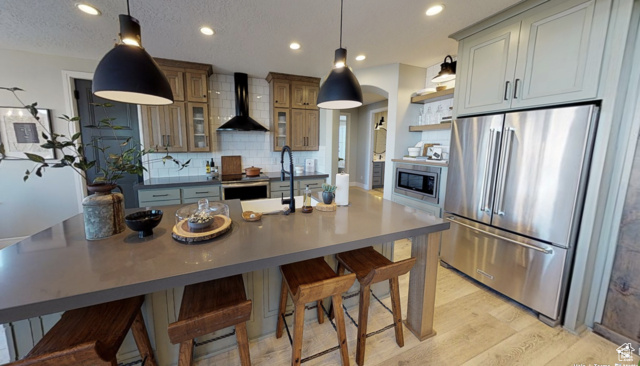 Kitchen with wall chimney exhaust hood, light hardwood / wood-style floors, a center island with sink, and appliances with stainless steel finishes