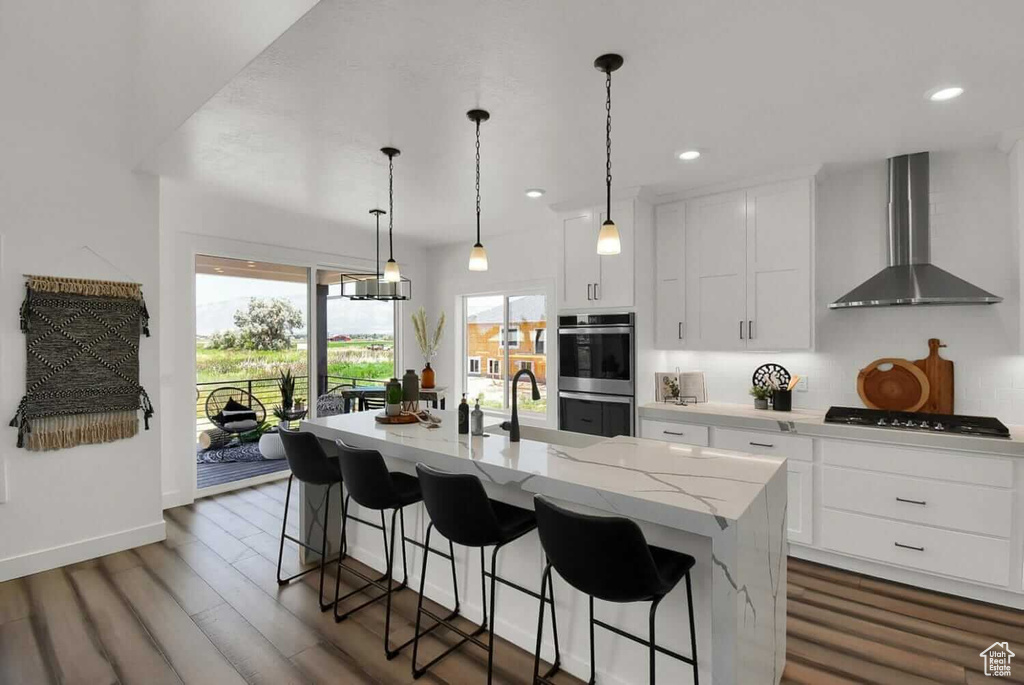 Kitchen with decorative light fixtures, wall chimney exhaust hood, an island with sink, dark wood-type flooring, and white cabinetry