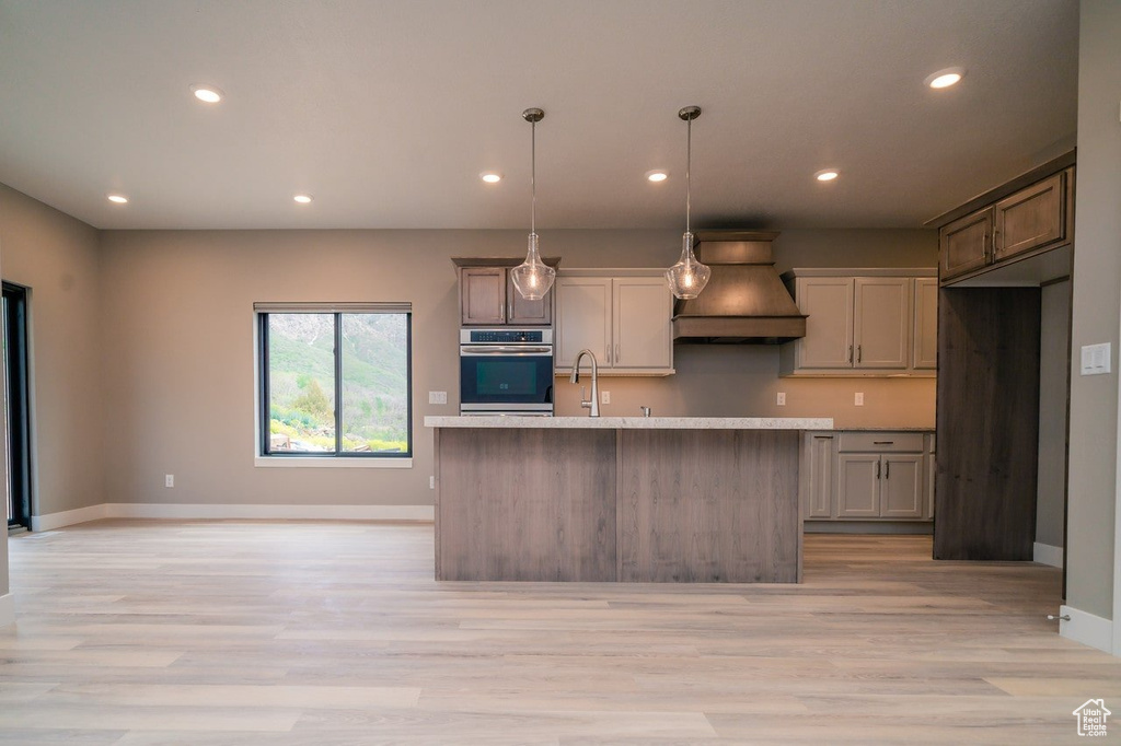 Kitchen with light hardwood / wood-style flooring, decorative light fixtures, premium range hood, an island with sink, and stainless steel oven