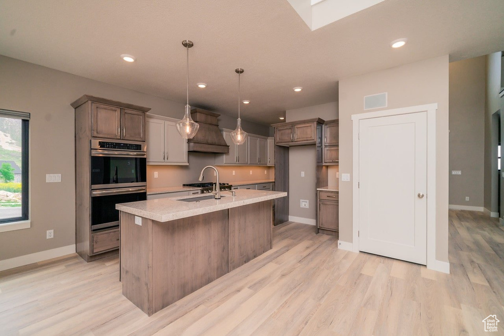 Kitchen with custom range hood, an island with sink, hanging light fixtures, stainless steel double oven, and light wood-type flooring