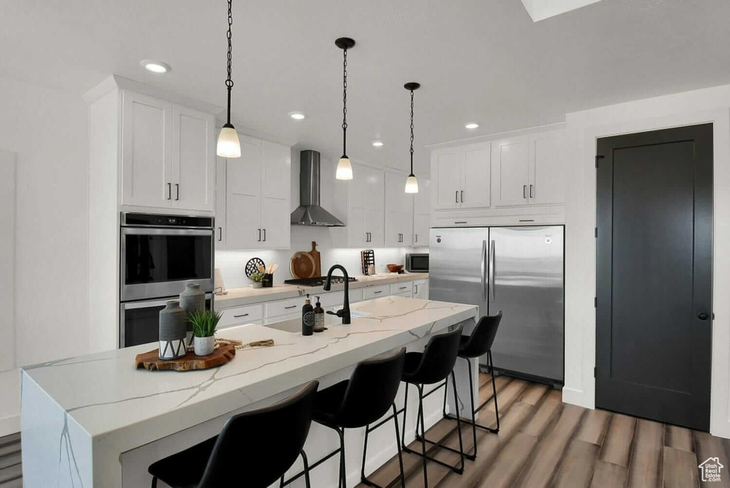 Kitchen with wall chimney range hood, white cabinets, stainless steel appliances, and a kitchen island with sink