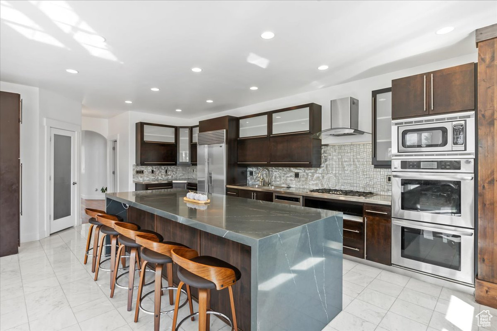 Kitchen with a kitchen island, built in appliances, a kitchen breakfast bar, wall chimney exhaust hood, and decorative backsplash