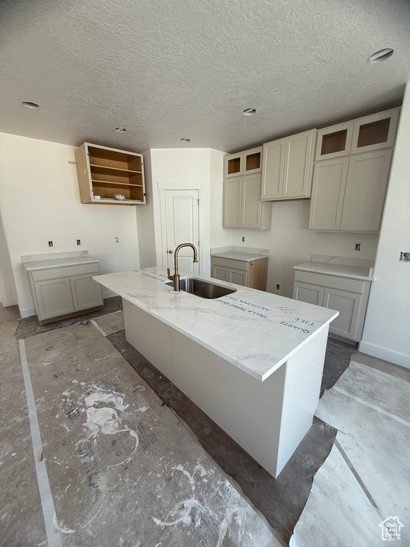 Kitchen with a sink, a center island with sink, a textured ceiling, baseboards, and light stone counters