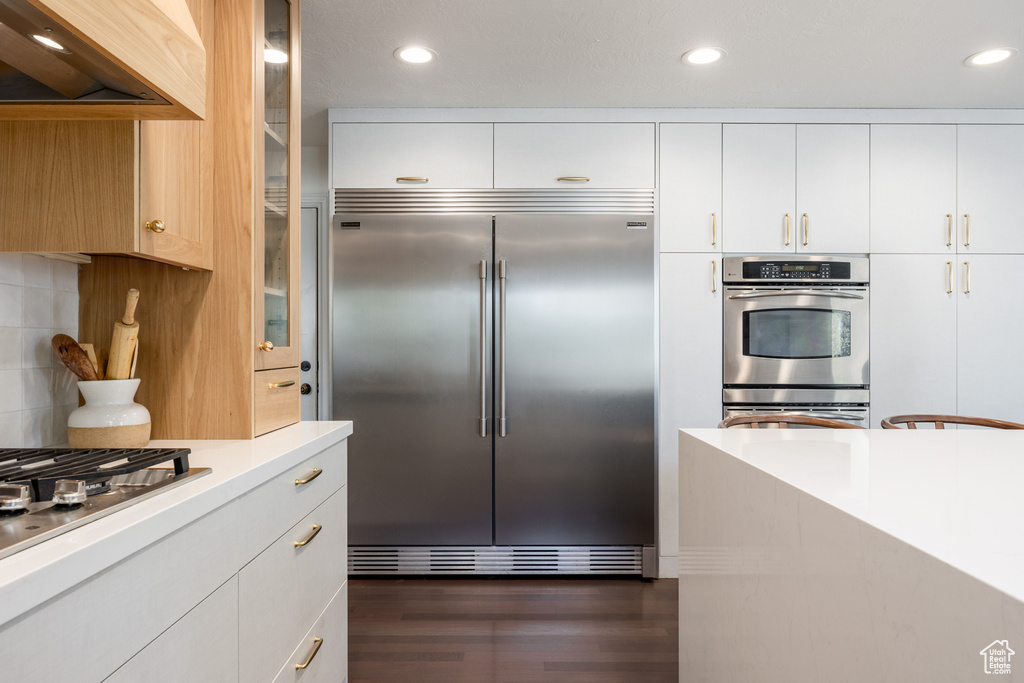 Kitchen with stainless steel appliances, custom exhaust hood, white cabinetry, dark wood-type flooring, and tasteful backsplash