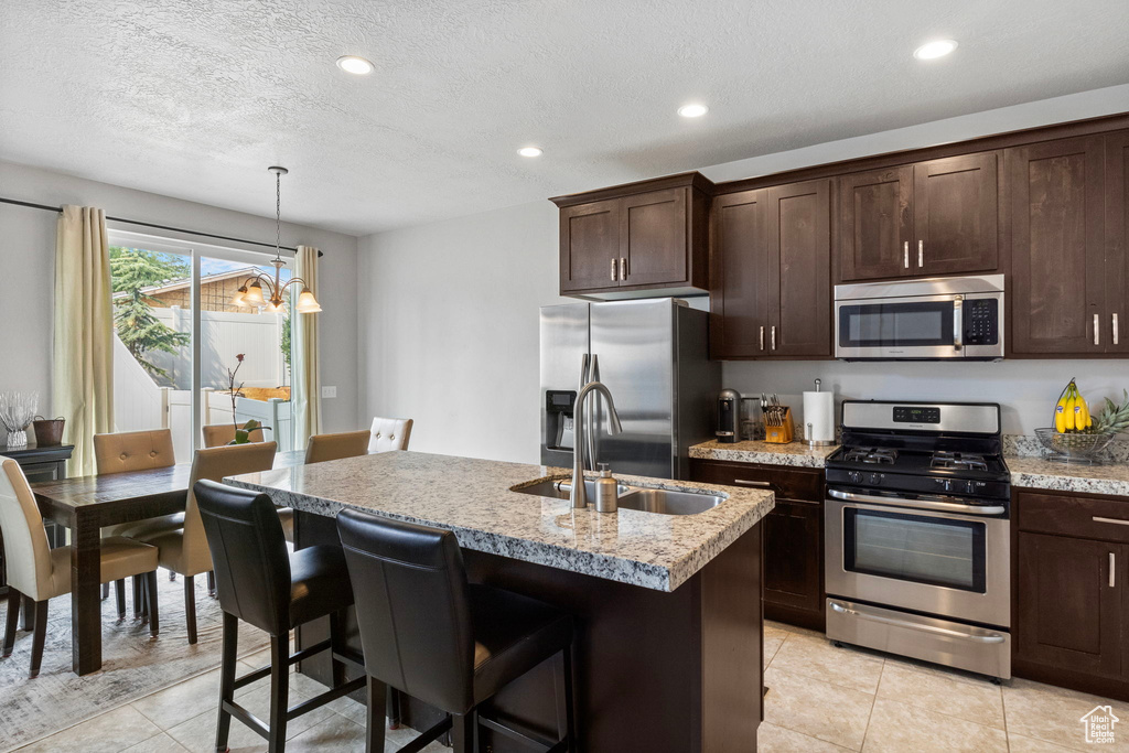 Kitchen featuring an inviting chandelier, appliances with stainless steel finishes, an island with sink, sink, and light stone counters