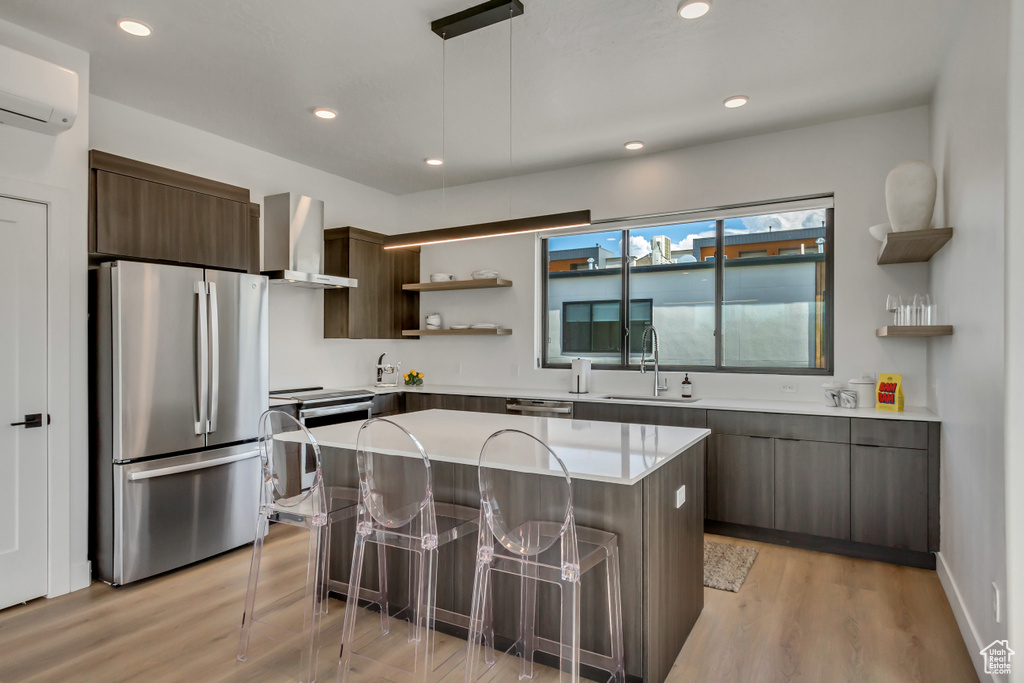 Kitchen featuring a center island, stainless steel appliances, wall chimney range hood, pendant lighting, and dark brown cabinetry