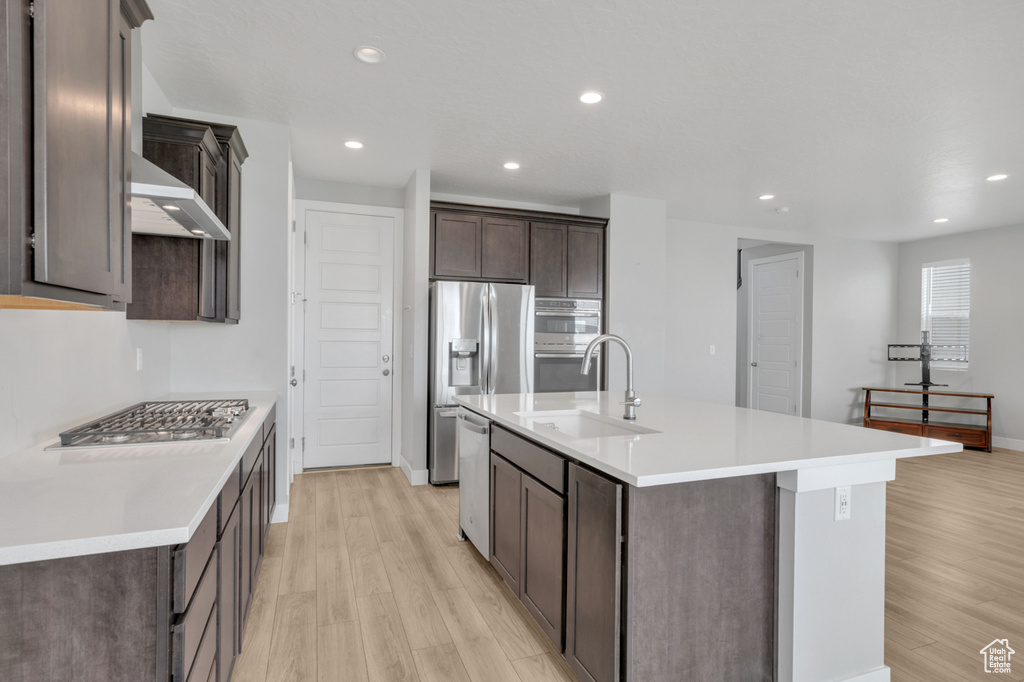 Kitchen with light wood-type flooring, stainless steel appliances, sink, a center island with sink, and dark brown cabinetry