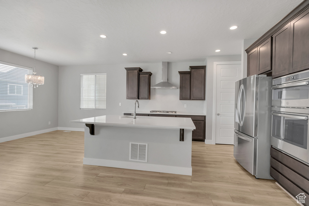 Kitchen featuring appliances with stainless steel finishes, light hardwood / wood-style floors, wall chimney range hood, dark brown cabinets, and a center island with sink