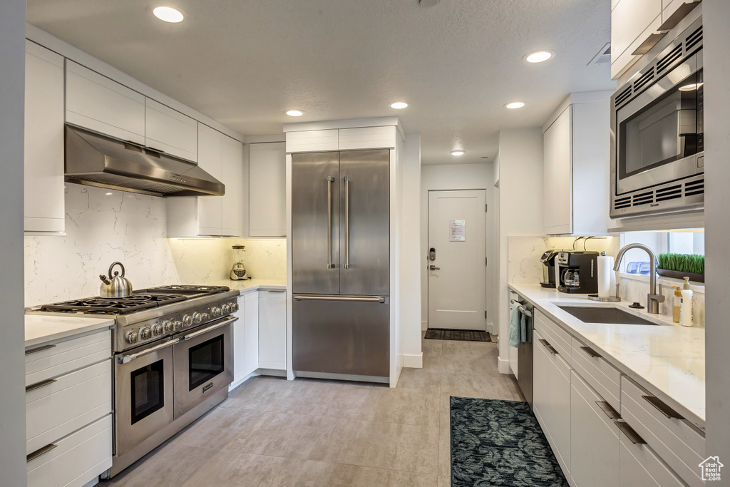 Kitchen with built in appliances, extractor fan, sink, white cabinetry, and light stone counters