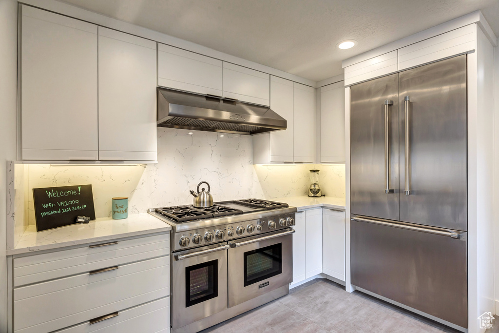 Kitchen featuring light stone countertops, range hood, tasteful backsplash, white cabinetry, and premium appliances