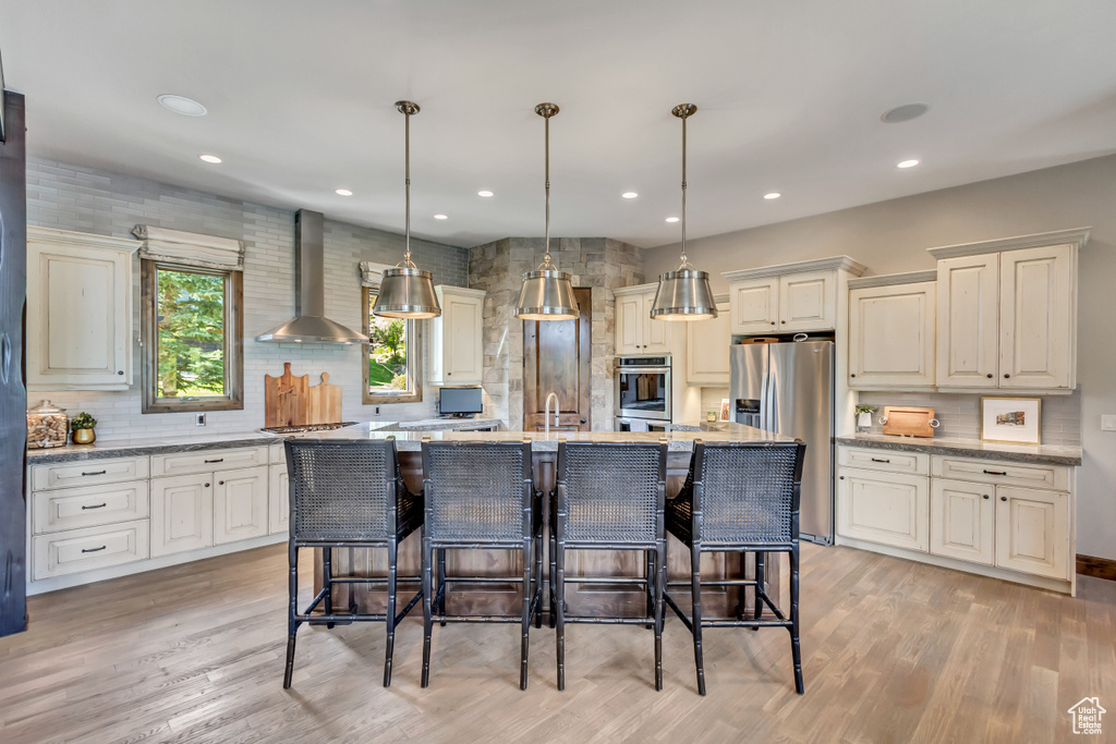 Kitchen featuring a center island with sink, light stone counters, appliances with stainless steel finishes, and wall chimney range hood