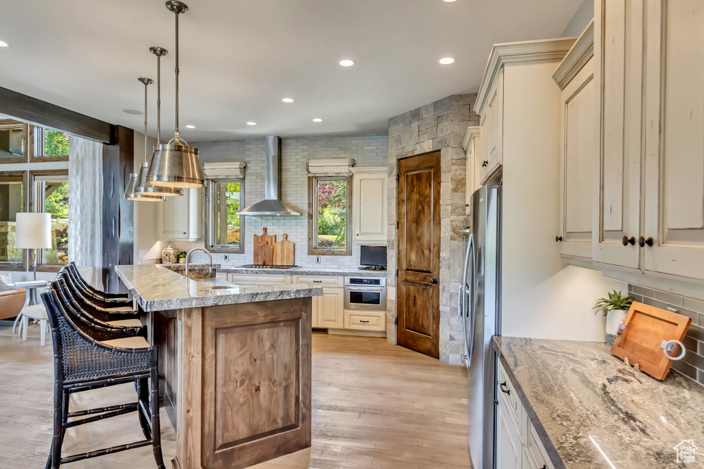 Kitchen featuring hanging light fixtures, appliances with stainless steel finishes, light stone countertops, wall chimney range hood, and a breakfast bar area