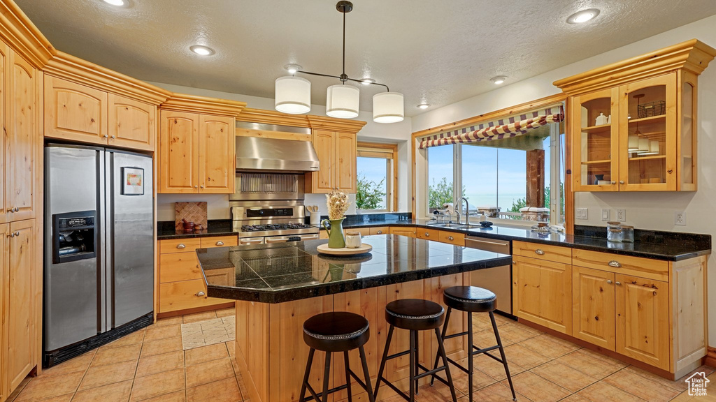 Kitchen with ventilation hood, light brown cabinetry, appliances with stainless steel finishes, a kitchen island, and a textured ceiling