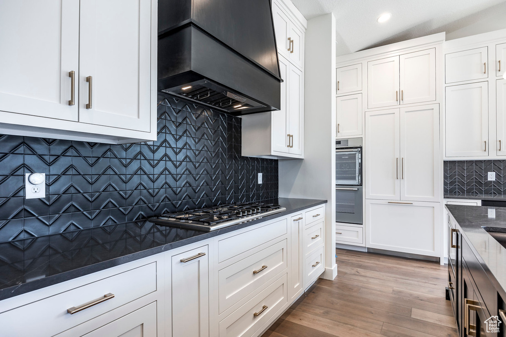 Kitchen with light wood-type flooring, decorative backsplash, wall chimney exhaust hood, black double oven, and white cabinets