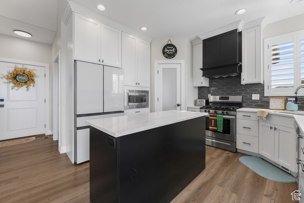 Kitchen with white cabinets, hardwood / wood-style flooring, custom range hood, appliances with stainless steel finishes, and a kitchen island