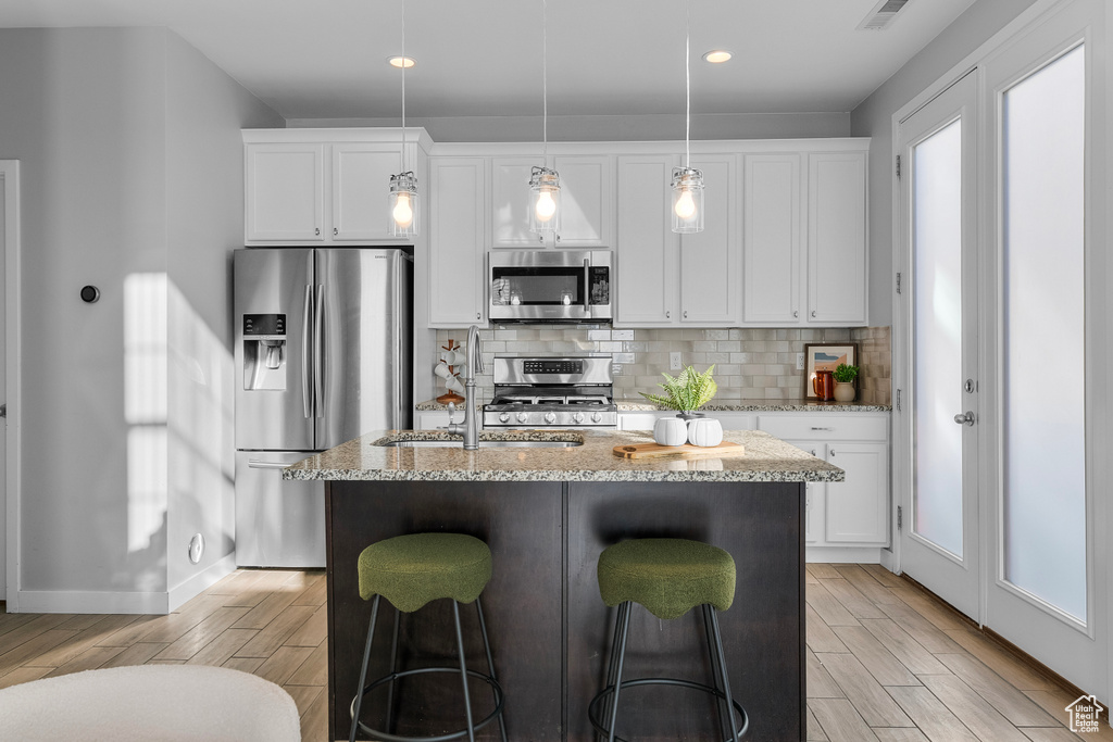 Kitchen with white cabinets, light stone counters, stainless steel appliances, light hardwood / wood-style floors, and a kitchen island with sink