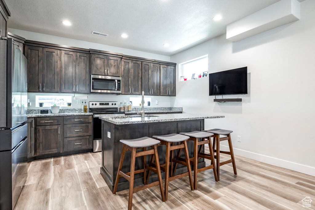 Kitchen featuring appliances with stainless steel finishes, a kitchen bar, a kitchen island with sink, and light stone counters