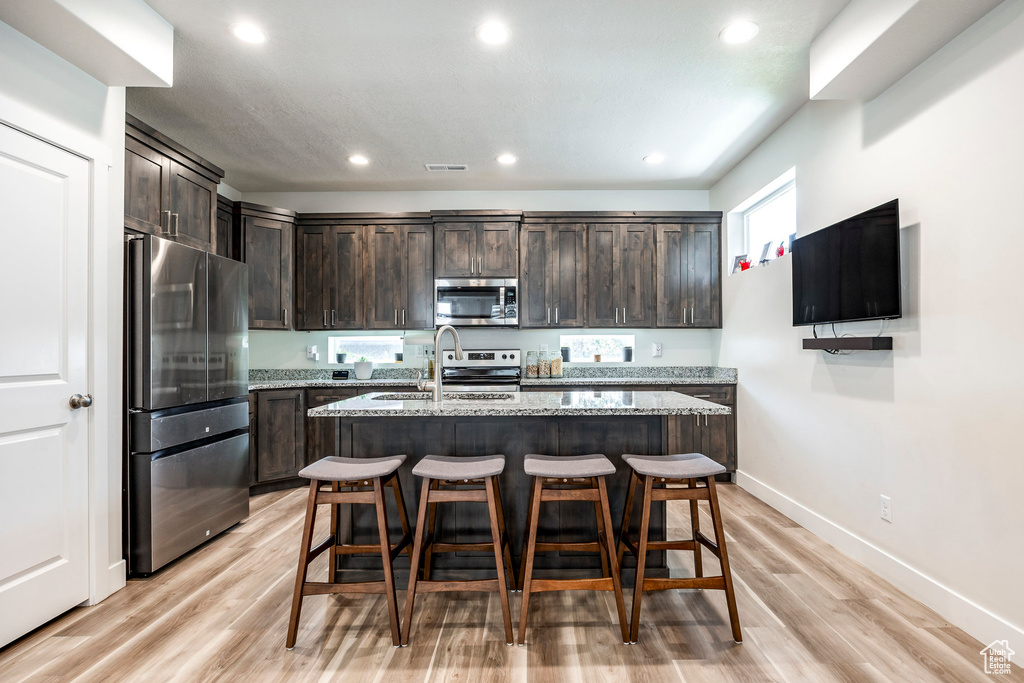 Kitchen with appliances with stainless steel finishes, dark brown cabinets, a sink, light stone countertops, and light wood finished floors