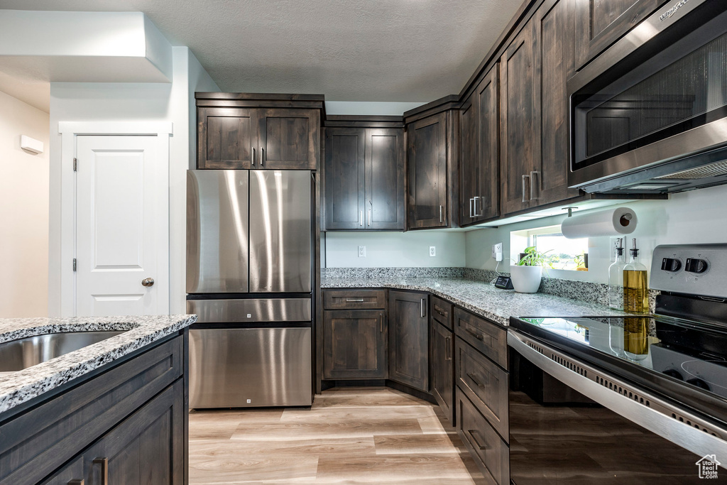 Kitchen with stainless steel appliances, dark brown cabinets, light stone countertops, light wood-style flooring, and a textured ceiling